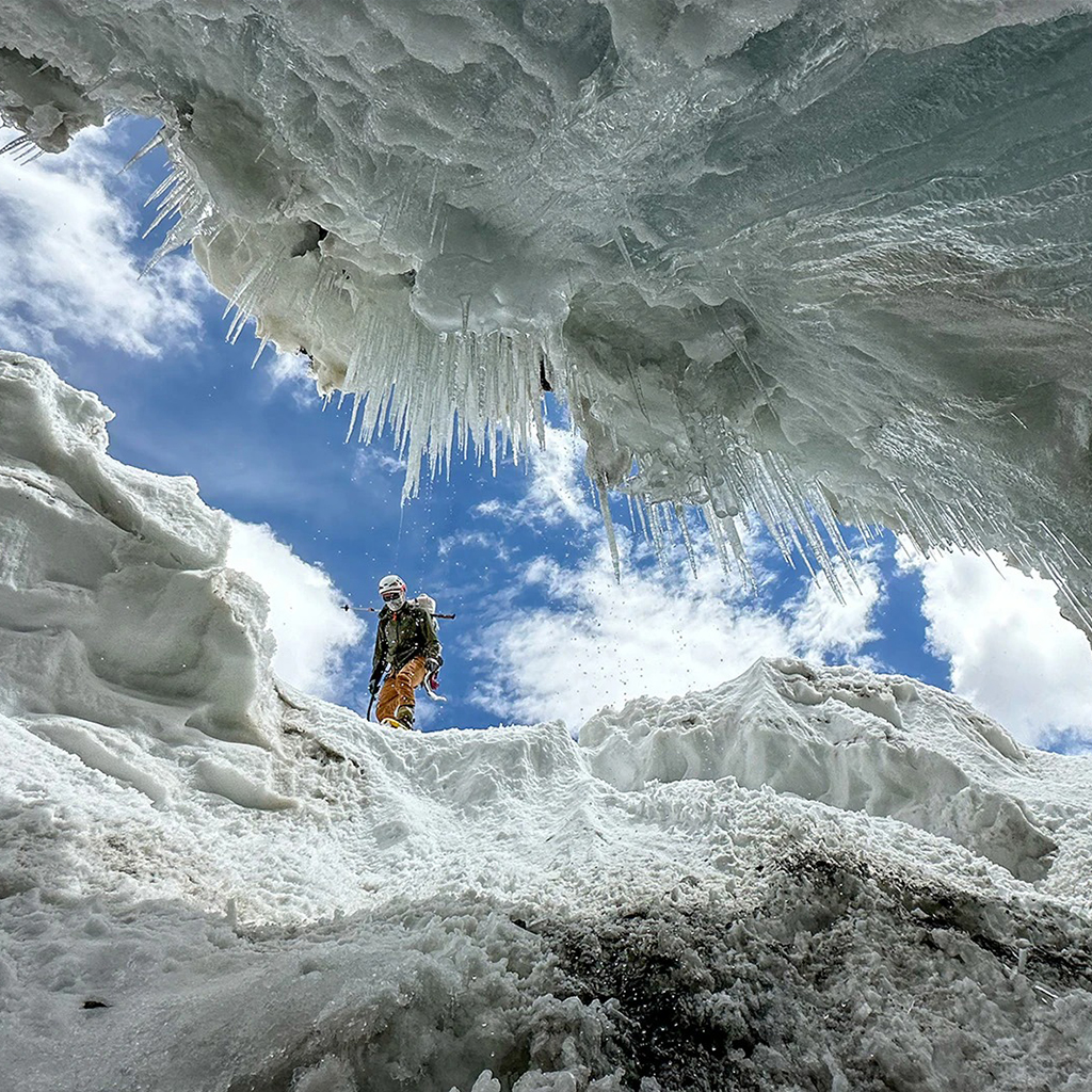 View from inside an icy cave looking up at a climber against a bright sky.