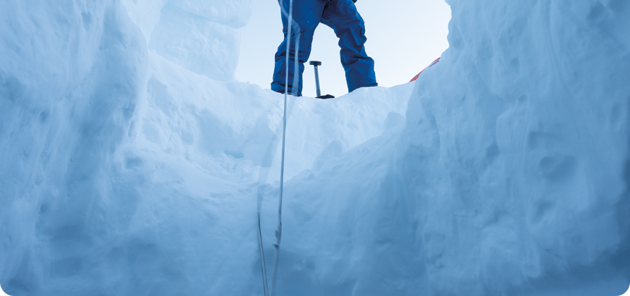 Person standing at the top of a snow cave opening with rope and winter gear