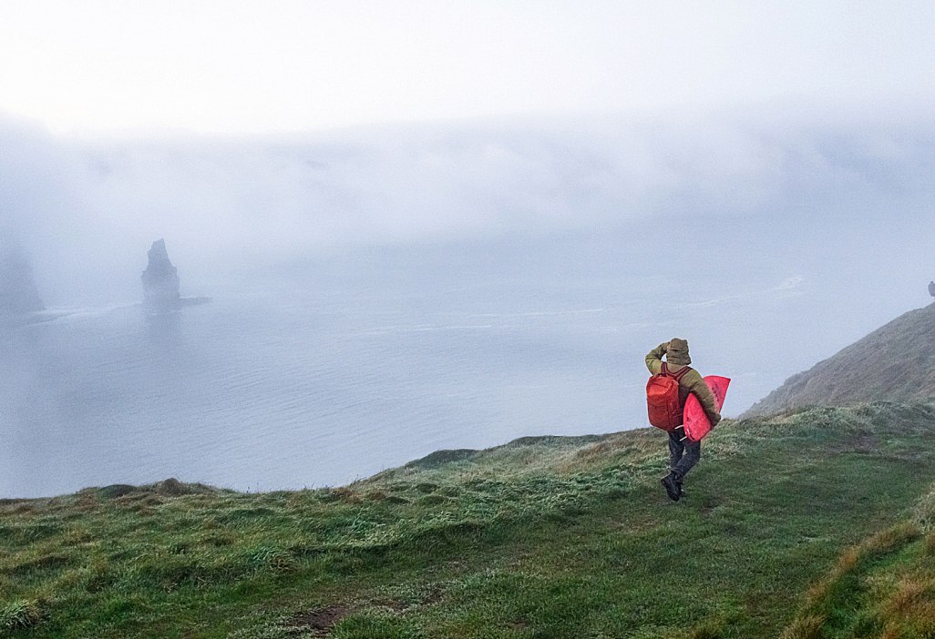 A person carrying a surfboard looks for wave along a foggy coastline.