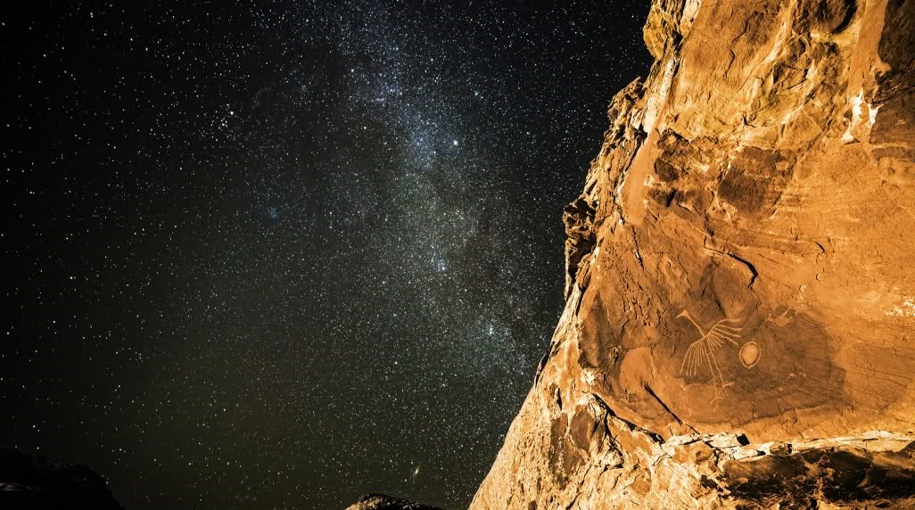 Stars in the night sky shine brightly above a petroglyph on a rock wall.