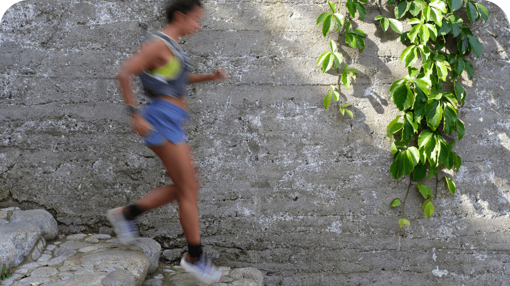 The blurry form of a runner wearing blue shorts and a yellow vest moves down stone steps. A green vine hangs from a stone wall next to her.